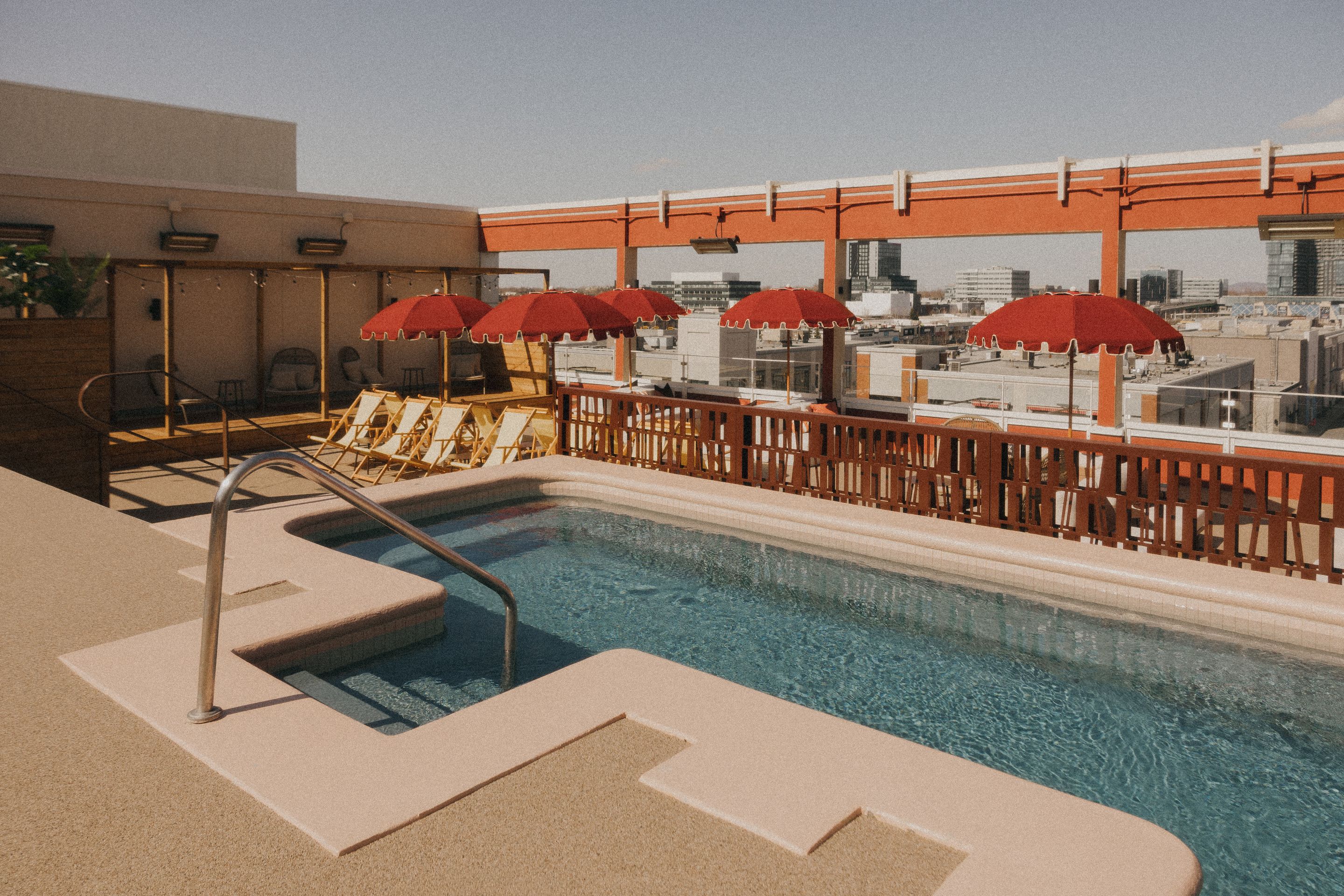 Piscine sur le toit d’AWŪ avec vue urbaine, mobilier extérieur et parasols rouges dans un espace consacré au bien-être.