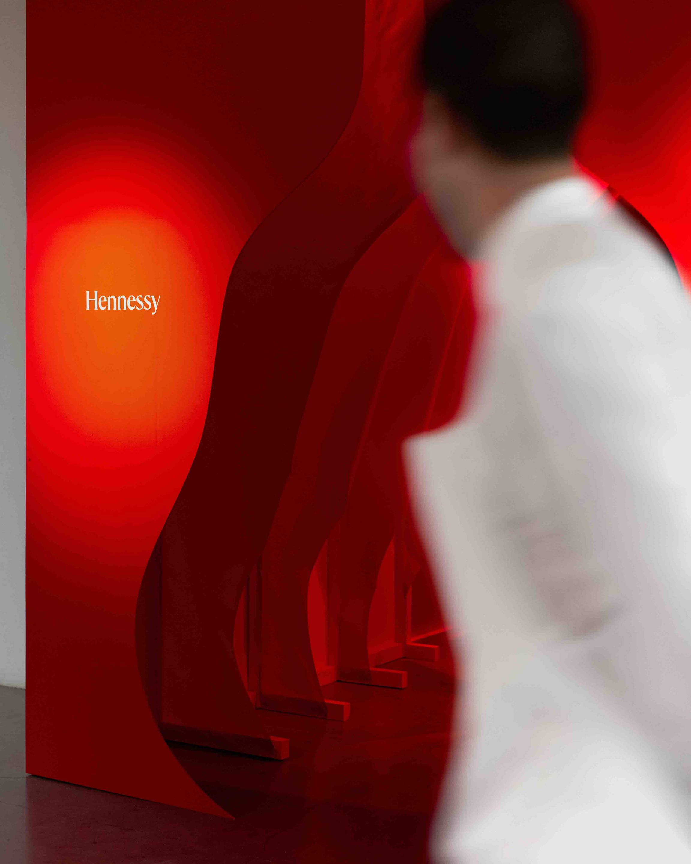 Installation scénographique rouge aux formes courbes évoquant une carafe Hennessy, avec un invité au premier plan.