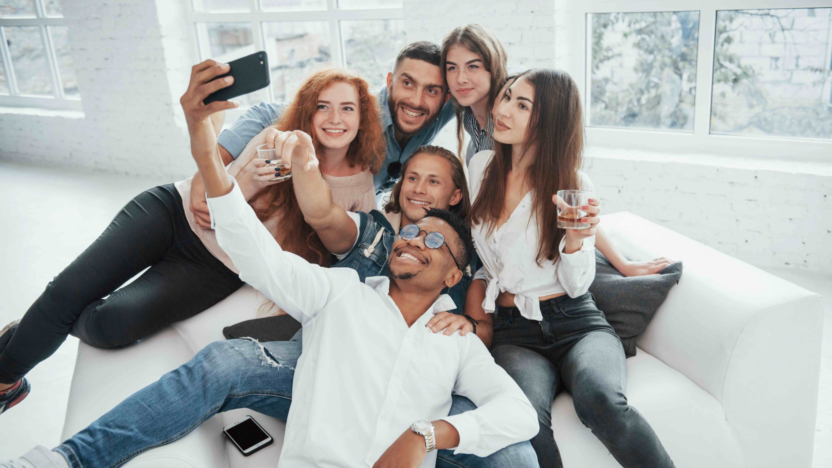 Groupe d’amis réunis dans un salon lumineux prenant un selfie lors d’un moment convivial à la maison.